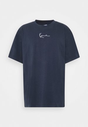 Person trägt rostfarbenes, geknöpftes Cordhemd über einem dunkelblauen T-Shirt mit Grafik, hellblaue hochgekrempelte Jeans, weiße Socken und weiße Sneaker mit lila Akzenten.
