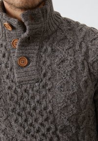 Maglione a trecce in lana grigio scuro con collo alto, caratterizzato da tre bottoni in legno marrone e un motivo testurizzato su tutto il tessuto.
