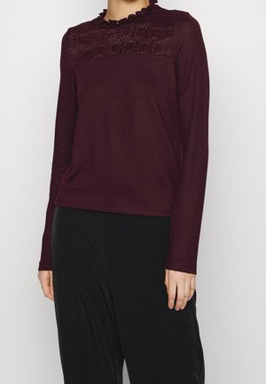 Femme portant un haut à manches longues bordeaux foncé avec détails en dentelle et col à volants, associé à un pantalon noir ample.