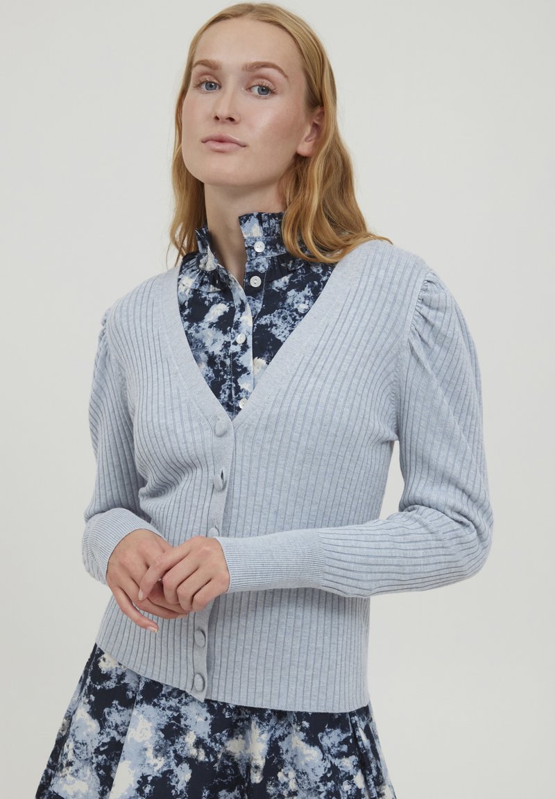 Cardigan en maille côtelée bleu clair avec un décolleté en V, une fermeture à boutons et des manches légèrement bouffantes, associé à une chemise à motif floral.