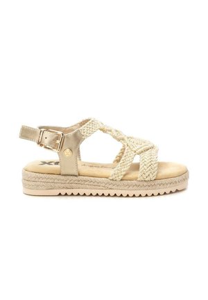 Sandalia plana beige con tiras de tela trenzada, una hebilla dorada en la correa del tobillo y una suela texturizada para mejor agarre.