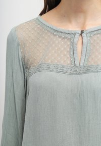 Blusa verde chiaro con una parte superiore in rete trasparente a pois, scollatura rotonda con apertura a occhiello e tessuto morbido e testurizzato.
