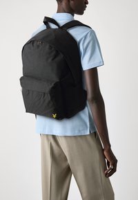 Un sac �à dos en tissu noir avec une texture lisse, doté d'une poche avant et d'un logo jaune subtil, porté sur le dos d'une personne vêtue de bleu clair.