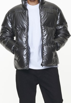 Personne portant une doudoune noire brillante par-dessus un t-shirt blanc et un pantalon noir, les mains dans les poches de la veste, debout devant un fond blanc.