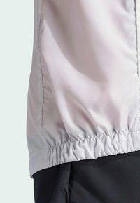 Giacca leggera di colore grigio pallido con un design testurizzato, orlo elasticizzato e finitura morbida, abbinata a tessuto nero sottostante.