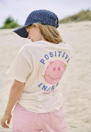 Jong persoon met een blauwe pet met hartjespatroon en een wit T-shirt met "Positive Energy" smiley ontwerp, staand op zanderig terrein.