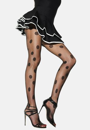 Jambes en collants noirs transparents à gros pois, portant des sandales à talons hauts noires et une jupe noire à volants bordés de blanc.