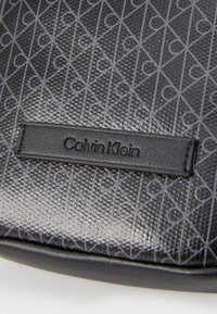 Fekete Calvin Klein táska, amelynek textúrája geometrikus mintázattal rendelkezik. Bőr logó címkével ellátva, amelyen bemélyített betűk találhatók.