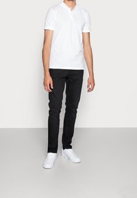Chemise polo blanche à manches courtes, associée à un jean noir ajusté et des baskets blanches. La tenue présente un design simple et épuré.