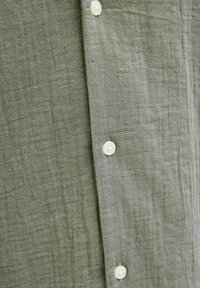 Olijfgroene linnen shirt met een gestructureerd oppervlak en transparante knopen langs de sluiting. Gekreukt stof voegt visuele interesse toe.