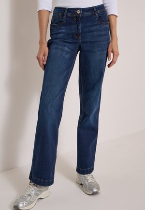 Femme portant un jean droit bleu foncé et des baskets argentées, debout devant un fond clair uni.