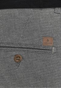 Tissu gris en gros plan montrant une poche arrière boutonnée et un patch en cuir marron marqué Jack & Jones.
