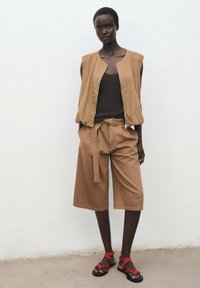 Beige ärmellose Weste über einem braunen gerippten Oberteil, kombiniert mit beigen weiten Shorts. Rote Sandalen vervollständigen das Outfit. Glattes Material und lässige Passform.