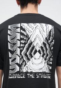 Μαύρο βαμβακερό μπλουζάκι με λευκό γραφικό τύπωμα στην πλάτη που απεικονίζει αφηρημένα σχήματα, ζιγκ-ζαγκ μοτίβα και το κείμενο "EMBRACE THE STRANGE".