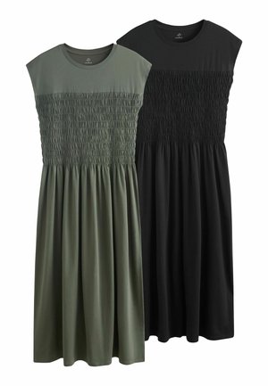 Frau steht an einer Wand und trägt ein ärmelloses, dunkelgrünes Maxikleid mit gerafftem Oberteil und schwarze flache Sandalen.