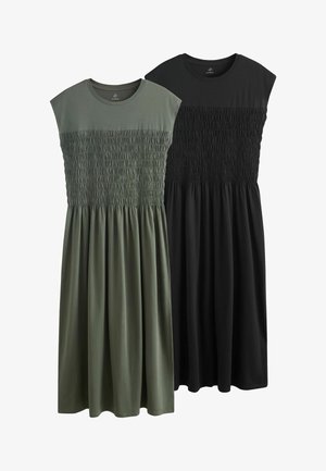 Donna in piedi contro un muro, indossa un vestito lungo verde scuro senza maniche con corpetto arricciato e sandali neri bassi.