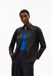 Black button-up shirt with a collar, long sleeves, and a relaxed fit. Worn over a blue crew neck top and paired with pinstriped trousers.