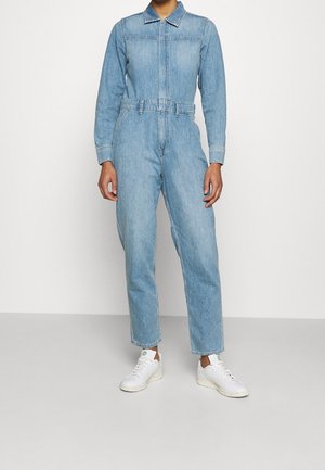 Combinaison en denim bleu clair à manches longues, avec col, fermeture éclair à l'avant et taille ceinturée. Stylée avec des baskets blanches et une coupe droite aux jambes.