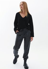 Schwarzer V-Ausschnitt-Pullover, lockerer Schnitt, gerippte Bündchen; kombiniert mit schwarzen, glänzenden, schmal zulaufenden Hosen mit elastischem Bund und Seitentaschen.