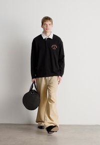 Μαύρο πουκάμισο πόλο με μακριά μανίκια και λευκό γιακά, χακί παντελόνι σε φαρδιά γραμμή και κυκλική μαύρη τσάντα. Μοντέλο που περπατάει σε ουδέτερο φόντο.