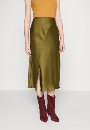 Kobieta w musztardowej bluzce bez rękawów, oliwkowo-zielonej sukience midi z satyny z przednim rozcięciem oraz bordowych zamszowych botkach do połowy łydki stojąca na białej podłodze.