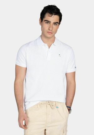 Joven de cabello oscuro que lleva una camisa polo blanca y pantalones beige con cordón, de pie con una mano en el bolsillo contra un fondo liso.