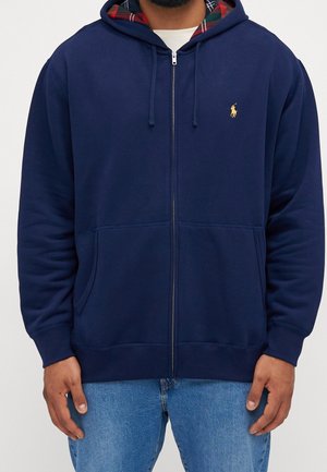 Homme portant un sweat à capuche zippé bleu marine avec capuche doublée en tissu écossais et logo brodé jaune, associé à un jean bleu clair.
