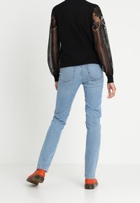 Zwarte trui met kantpofmouwen, lichtblauwe rechte jeans en oranje schoeisel, gezien vanaf de achterkant tegen een witte achtergrond.