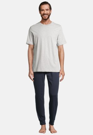 Man staat op blote voeten, glimlachend, draagt een lichtgrijs T-shirt met korte mouwen en donkerblauwe comfortabele broek tegen een effen achtergrond.