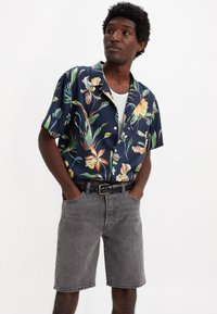 Kurzärmliges marineblaues Blumenshirt mit lebendigen grünen und orangefarbenen Mustern, kombiniert mit grauen Jeansshorts und einem geflochtenen schwarzen Gürtel.