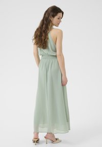 Light green, sleeveless maxi dress with a smocked waist, flowing fabric, and a subtle sheen. Worn with light-colored heeled sandals.