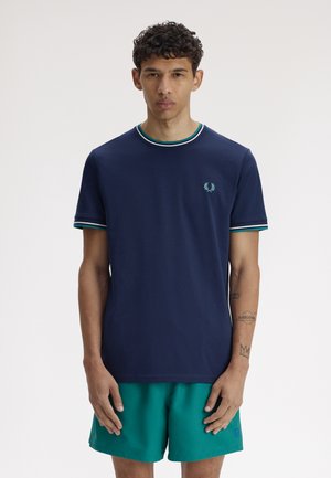 Joven vestido con una camiseta azul marino con cuello y mangas a rayas, y pantalones cortos color verde azulado, posando contra un fondo liso.