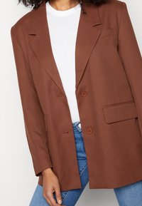 Mujer vistiendo un blazer marrón con botones sobre una camisa blanca y jeans azules, posando con la mano apoyada en el muslo.