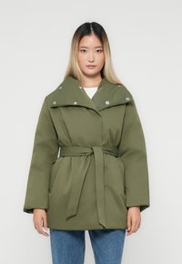 Kvinna klädd i en grön belagd vadderad jacka med tryckknappar på kragen, i kombination med blåjeans, stående mot en enfärgad vit bakgrund.