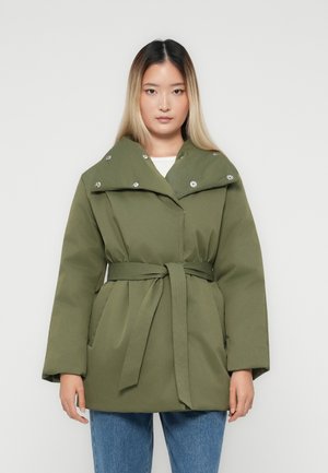 Kobieta w zielonej kurtce o pikowanej fakturze z paskiem w talii i napami przy kołnierzu, w połączeniu z niebieskimi dżinsami, stojąca na tle białego tła.