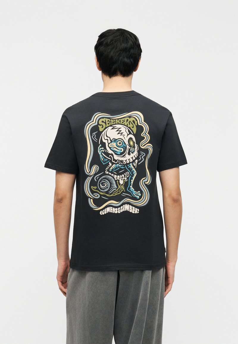 Άτομο που φοράει μαύρο μπλουζάκι με πολύχρωμο γραφικό μιας μπλε δημιουργίας μέσα σε κρανίο και τη λέξη "Seekers" στην πλάτη.