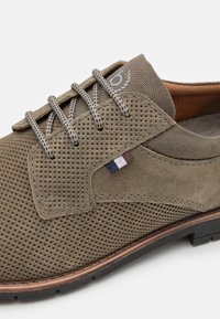 Zapato de gamuza perforada verde oliva con cordones grises. Presenta una pequeña etiqueta de color, un acento de cuero marrón en el talón y una suela de goma negra.