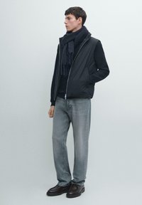 Czarna ocieplana kamizelka na ciemnym swetrze, szare dżinsy i ciemne buty. Model nosi teksturowany ciemny szalik z frędzlami. Prosty, nowoczesny design.