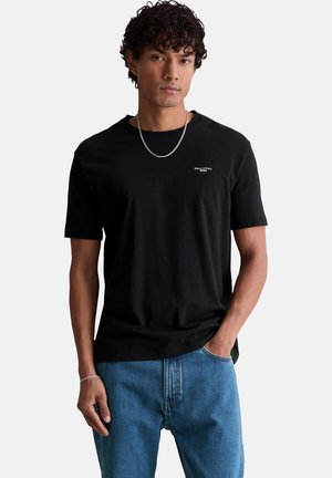Junger Mann mit lockigem Haar, der ein schwarzes Marc O'Polo T-Shirt, blaue Jeans und eine silberne Ketten-Halskette vor einem einfarbigen Hintergrund trägt.