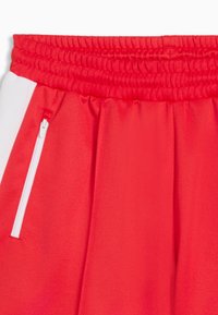 Shorts de sport rouges avec taille élastique et poche latérale zippée blanche, ornés d'une bande verticale blanche sur le côté.