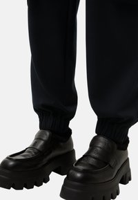Zwarte dikke platform loafers met gekorreld leer en een "B" logo, gedragen met zwarte broek met elastische manchet tegen een effen achtergrond.