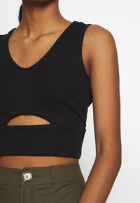 Haut de tank top noir raccourci en tissu côtelé, avec un décolleté en V profond et un détail découpé à la taille. Associé à un pantalon vert olive.