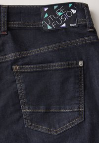 Vaqueros de mezclilla oscura con costuras amarillas, un bolsillo trasero con botón y una etiqueta de marca con un diseño geométrico.