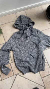 Grey knit hoodie with buttons and drawstrings lying flat on beige tiled floor.