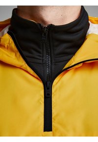 Žlutá bunda s černou vnitřní vrstvou, vybavená zipem a vysokým límcem. Materiál působí hladce a lehce.