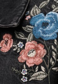 Μπλε, ροζ και λευκή κεντητική με λουλούδια σε σκούρο τζιν ύφασμα με εμφανείς λεπτομερείς ραφές και διπλωμένη άκρη.