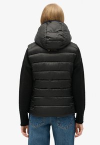 Gilet matelassé noir avec capuche, présentant un design matelassé, une texture douce et un accent logo à l'arrière, associé à un haut noir à manches longues.
