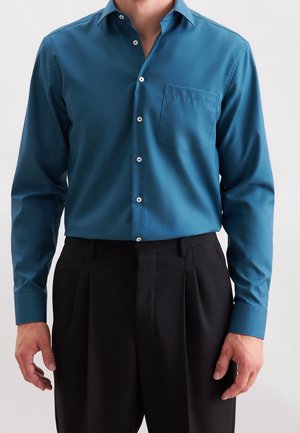 Man draagt een teal gekleurde blouse met lange mouwen en knopen, voorzien van een borstzakje, en zwarte geplooide broek, staand tegen een effen achtergrond.