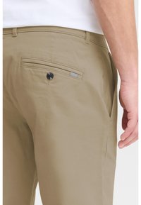 Homme portant un pantalon beige avec une poche arrière boutonnée et une chemise blanche, main détendue le long du corps sur un fond uni.