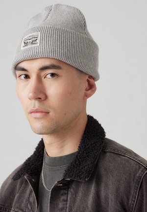 Gray ribbed knit beanie with a folded cuff, featuring a label patch. Worn with a black denim jacket, showing a soft black fur collar.